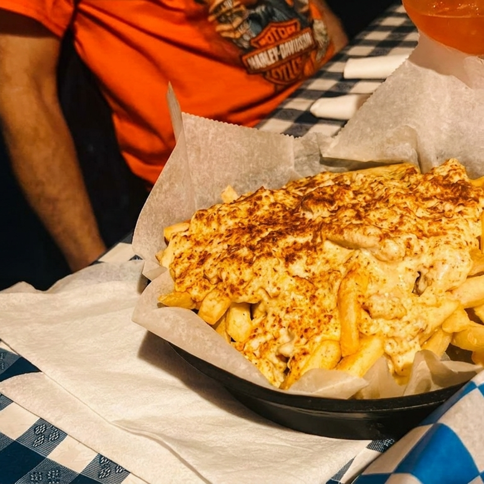 A basket of cheesy fries with a person in an orange shirt and drinks in the background.