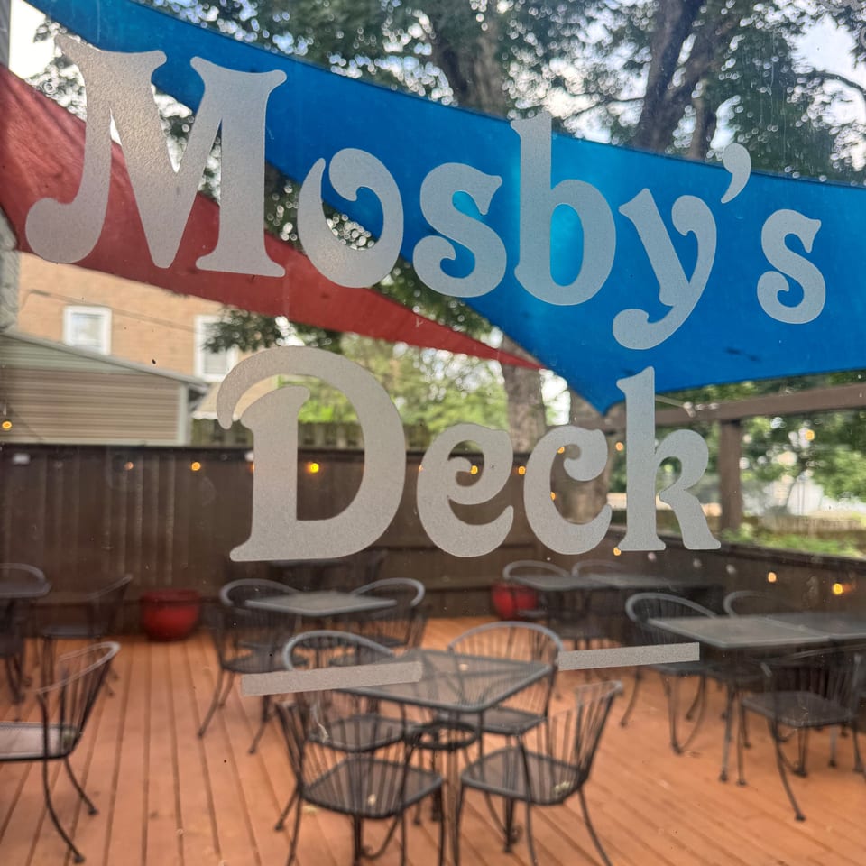 Restaurant window lettering overlooks an outdoor wooden patio furnished with black wire tables and chairs.