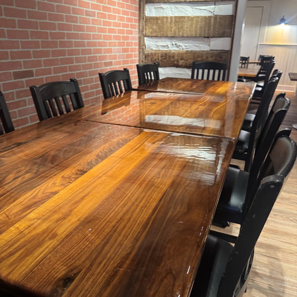 Rustic restaurant dining room with a long polished wooden table, black chairs, brick walls, and exposed wood accents.