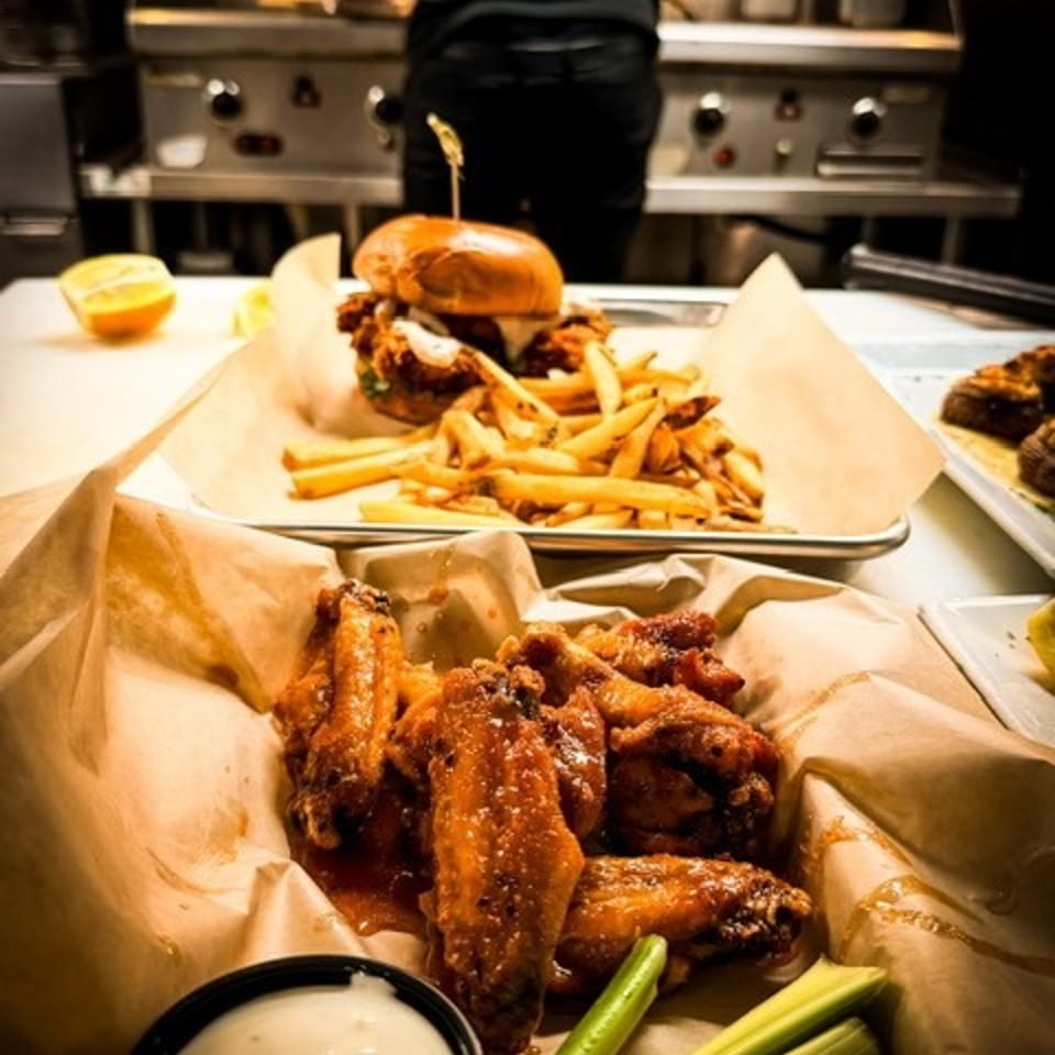 Restaurant counter displays crispy chicken wings with celery and dipping sauce, alongside a burger and fries in a busy kitchen.