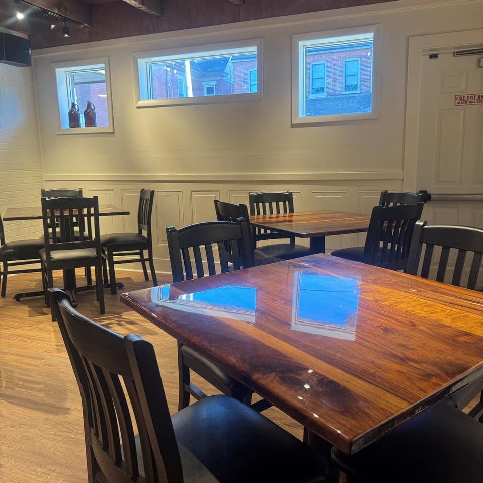 Restaurant dining room with polished wooden tables, black chairs, exposed timber ceiling beams, and windows overlooking brick buildings.