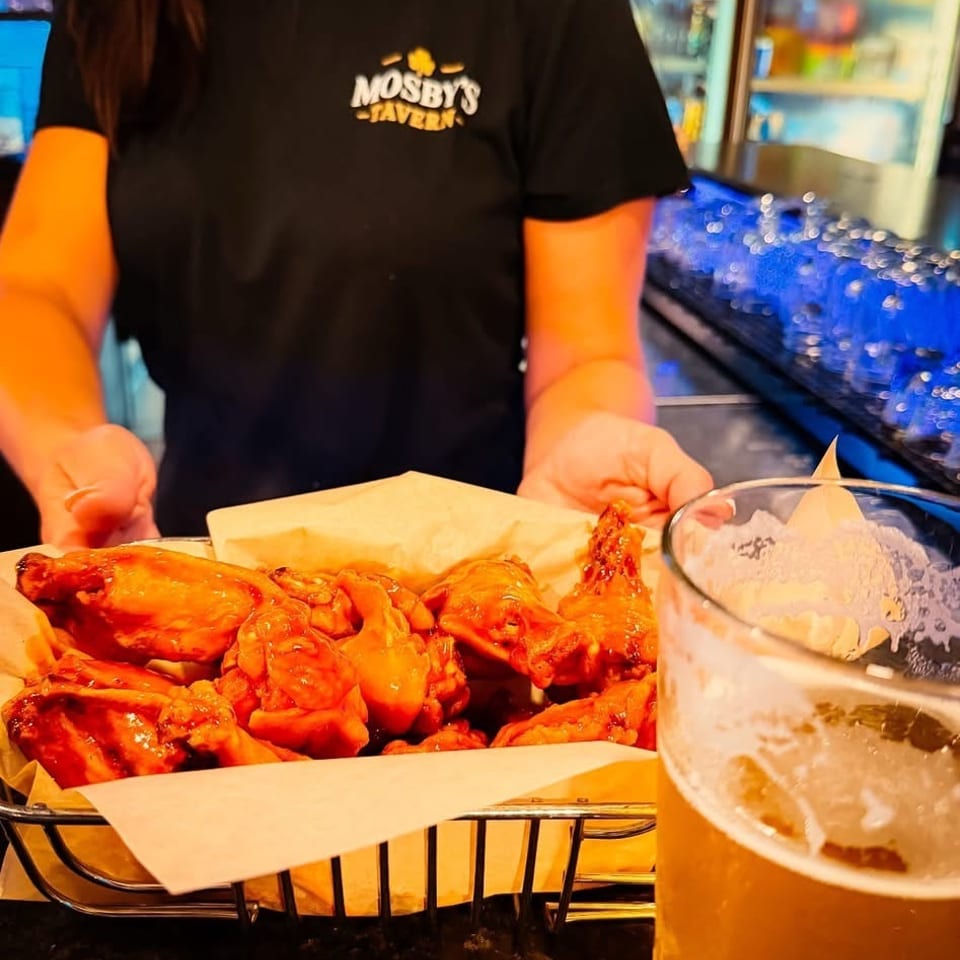 Restaurant server presents saucy chicken wings beside a large iced drink, with beverage coolers in the background.